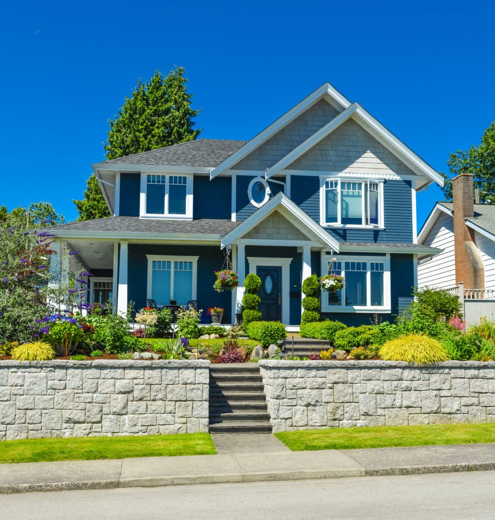 Large blue two-story house with a stone retaining wall and manicured garden.