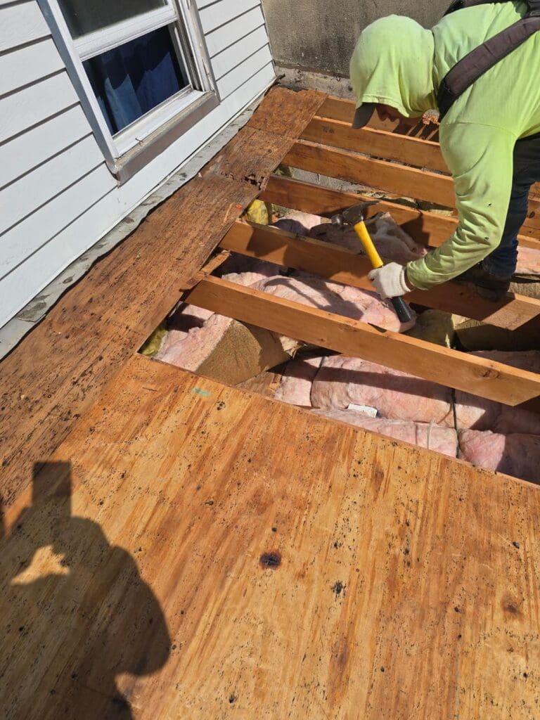 Worker in a lime hoodie hammers apart a wooden deck, exposing pink insulation between joists beside a house.